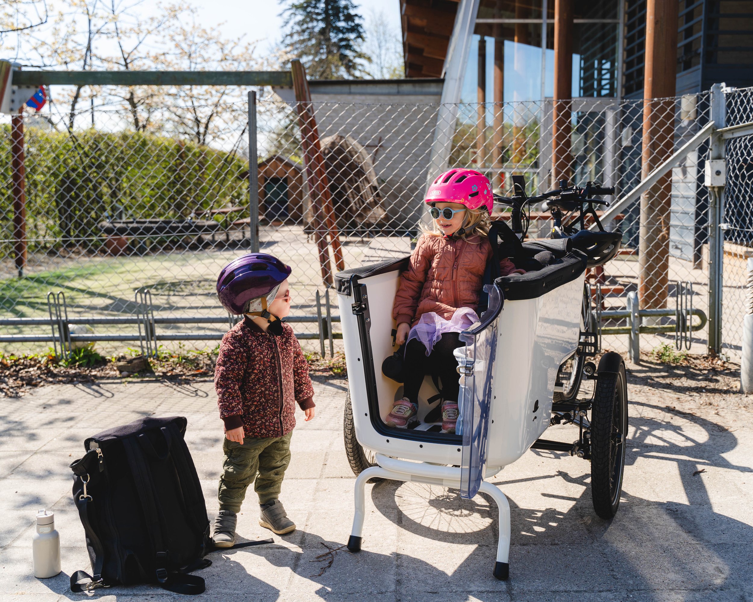 Zwei Kinder mit Fahrradhelmen. Ein Kind sitzt in einem Lastenfahrrad, das andere steht daneben. Im Hintergrund ist ein Zaun und ein Gebäude. Draußen scheint die Sonne, eine Tasche und eine Flasche stehen auf dem Boden.