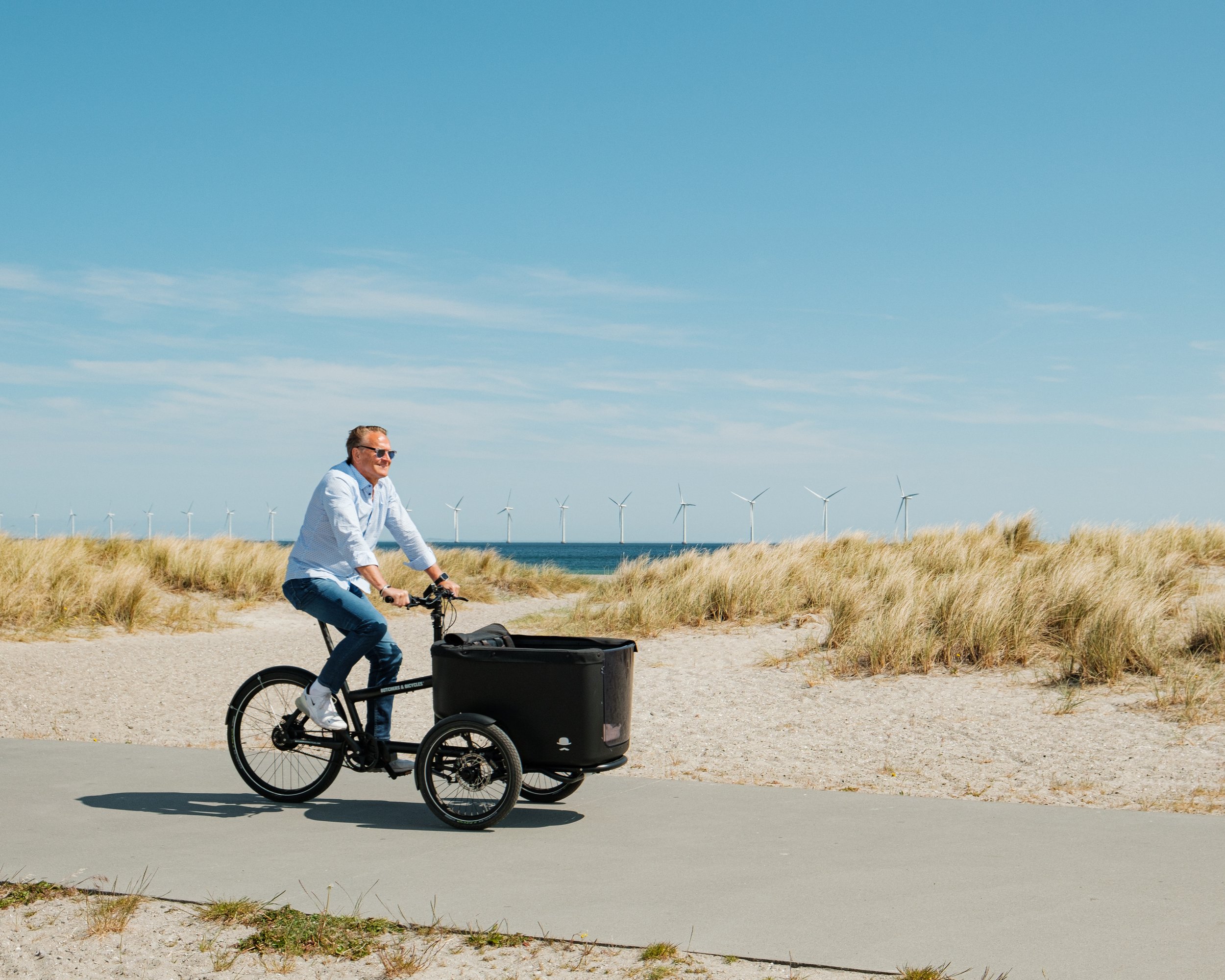 Mann fährt ein Lastenfahrrad auf einem Weg am Strand mit Windkraftanlagen im Hintergrund unter klarem blauem Himmel.