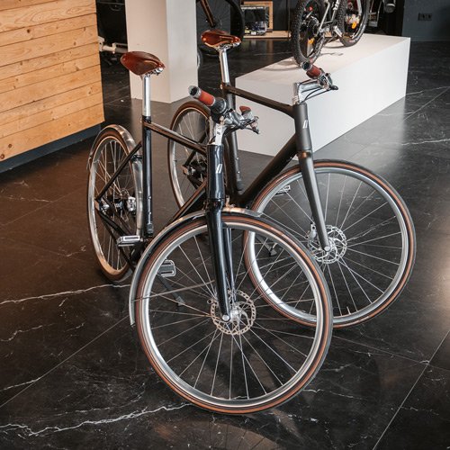 Zwei moderne Fahrräder mit schwarzen Rahmen stehen auf einem dunklen Marmorboden in einem Geschäft oder Showroom, umgeben von Holzwänden und anderen Fahrrädern.