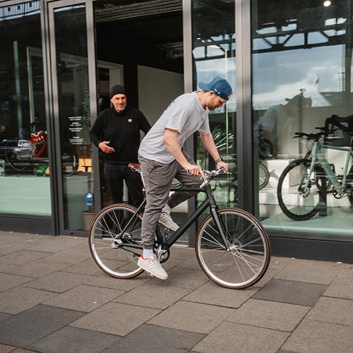 Ein Mann in einem grauen T-Shirt und blauer Mütze fährt ein schwarzes Fahrrad auf dem Bürgersteig vor einem Gebäude mit großen Fenstern. Eine andere Person steht im Hintergrund in schwarzer Kleidung.