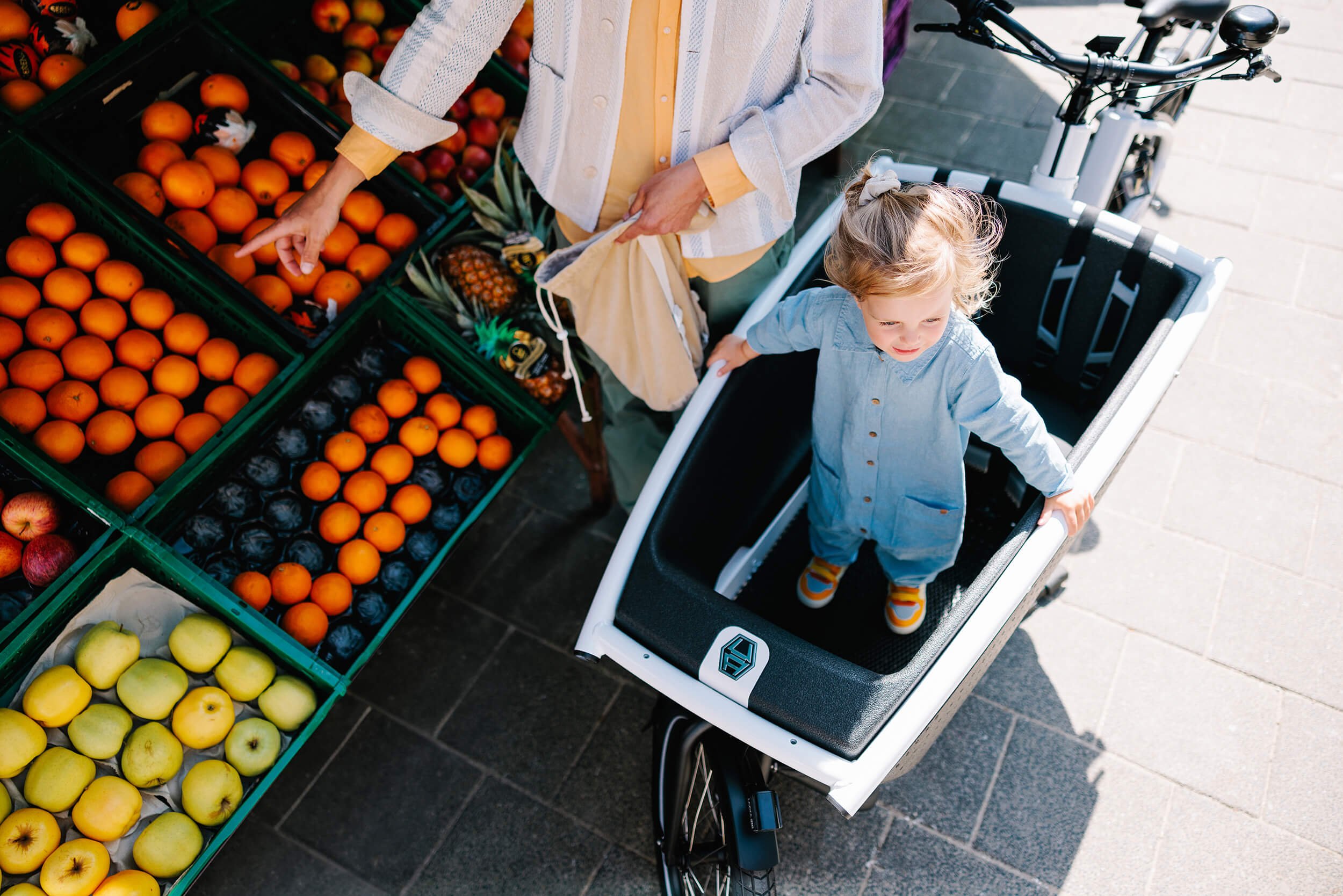 Person kauft Obst an einem Marktstand; Kind steht in einer Fahrradkindersitz-Kiste.