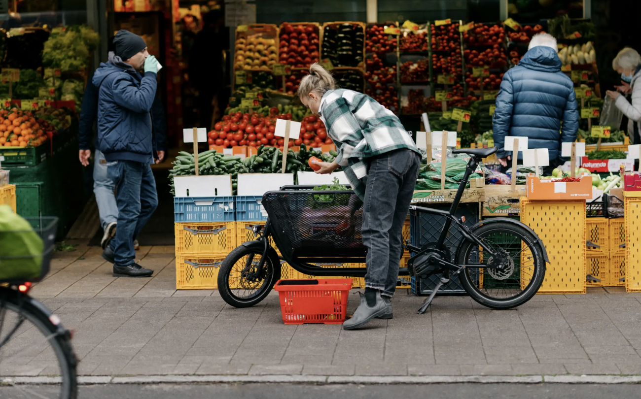 Personen und Gemüsemarkt im Freien, leuchtend gefüllte Obst- und Gemüsekisten, eine Person mit Fahrrad und Einkaufskorb im Vordergrund.